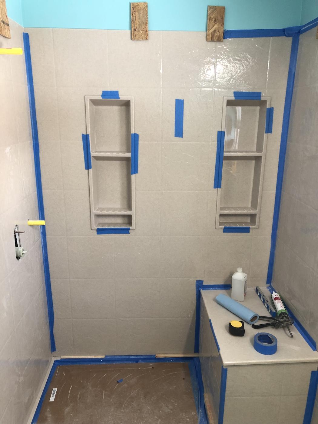 The shower mid-installation: wall panels braced with timber offcuts, painter's tape along every edge, protective film still on, a caulk gun and a level resting on the bench